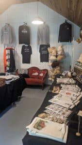 Back wall of the gift shop at Maple Hill Manor featuring sweatshirts hanging on backwall. Tea towels on the table to the right.